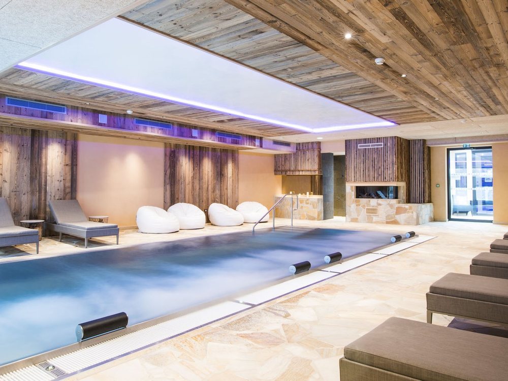 La piscine intérieure de l'Hostellerie des Châteaux, plafond de vieux bois, transats et poufs blancs, cheminée au fond