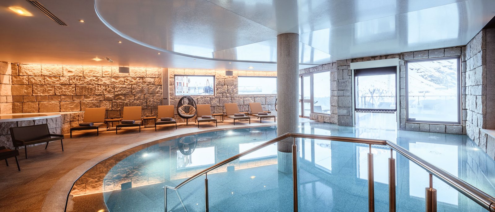 La piscine intérieure de l'Altapura, son bassin arrondi entre murs de pierre et baies vitrées ouvertes sur la neige