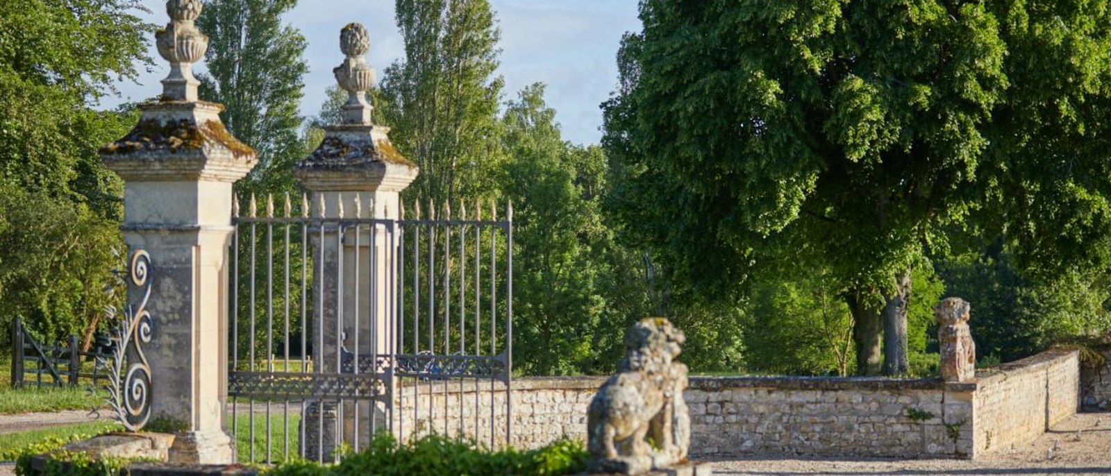 La grille d'honneur du Château d'Audrieu, piliers de pierre à pots à feu, lions sculptés et mur d'enceinte sous les grands arbres