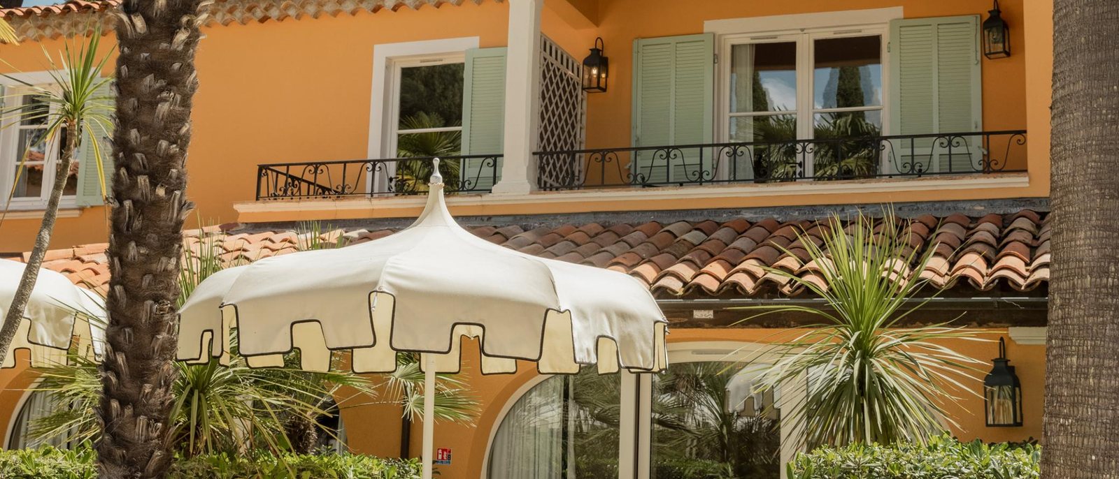 Un mas provençal de La Bastide de Saint-Tropez, façade ocre et volets vert d'eau, derrière un parasol blanc à franges et des palmiers