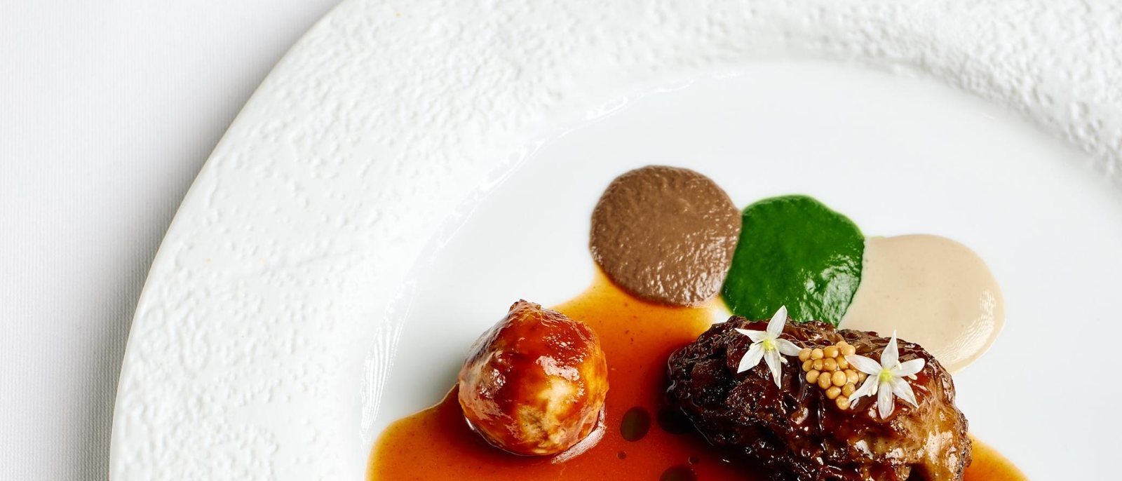 Une assiette du Jardin de la Bastide, deux pièces glacées, une sauce ambrée, des points de purée verte et brune et des fleurs blanches