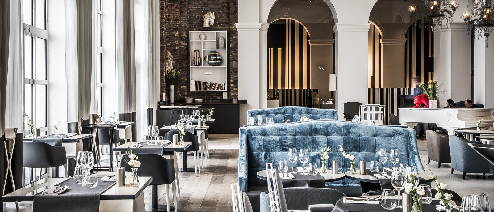 La salle du restaurant La Galerie, ses tables dressées, sa banquette de velours bleu, ses arcades blanches et ses grandes fenêtres