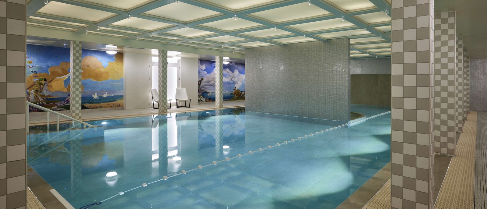 La piscine d'eau de mer des Cures Marines, sous un plafond à caissons lumineux, entre des colonnes à damier et de grandes fresques de bord de mer