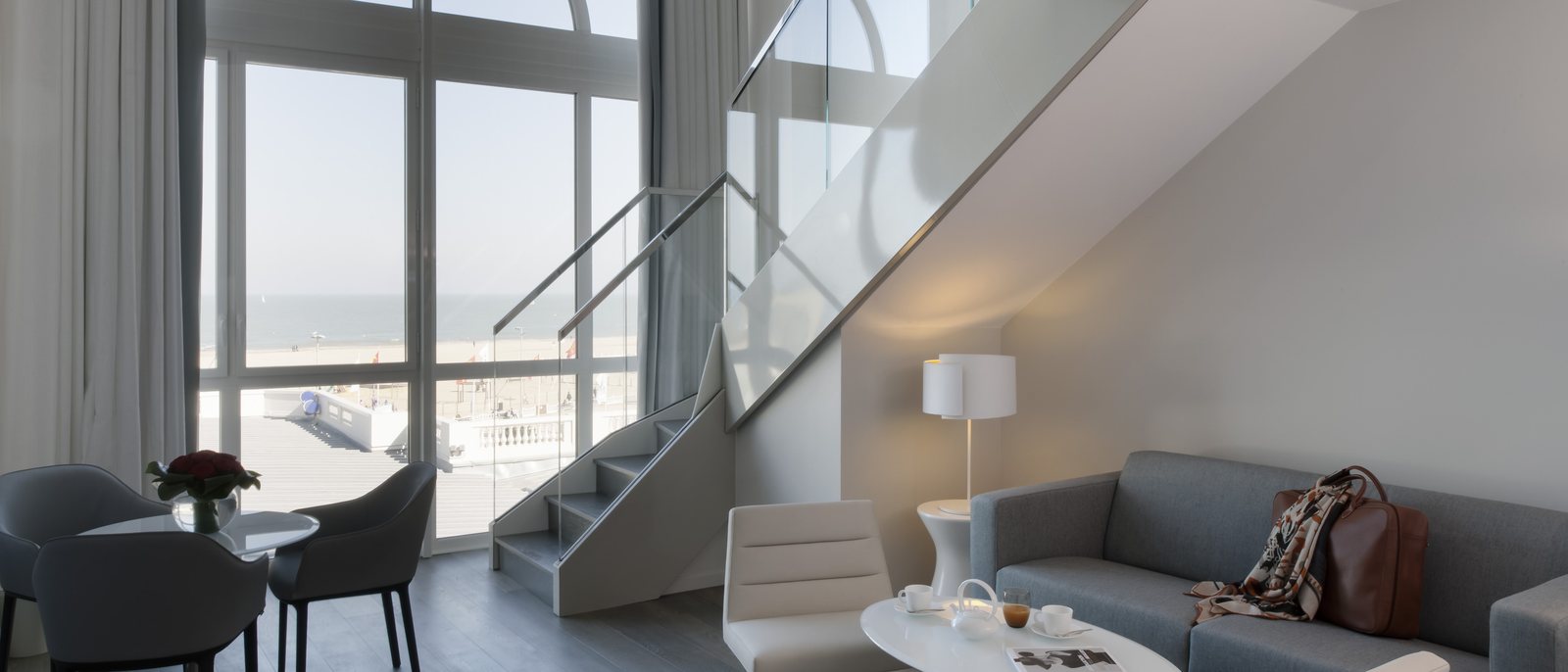 Le salon en duplex de la suite Reine des Plages, sa grande baie sur la plage de Trouville, son canapé gris et son escalier