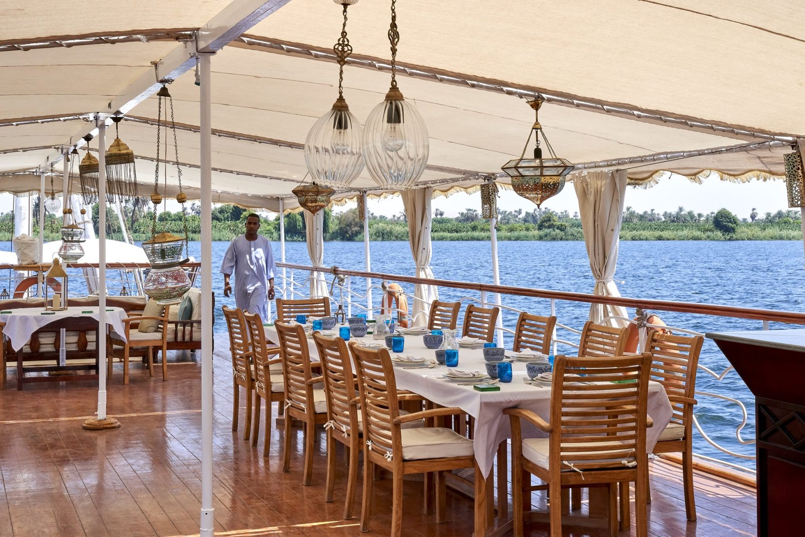 Table dressée sous le taud du pont supérieur d'une dahabieh Lazuli, lanternes suspendues et rive du Nil