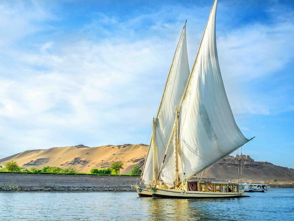 Une dahabieh à deux voiles latines devant les dunes et la rive rocheuse d'Assouan