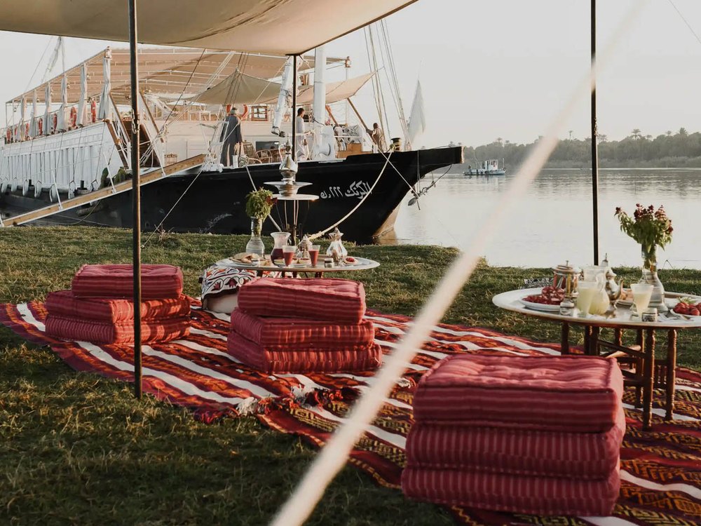 Dîner dressé sur la berge du Nil sous un taud de toile, tapis, coussins de sol et dahabieh amarrée au crépuscule