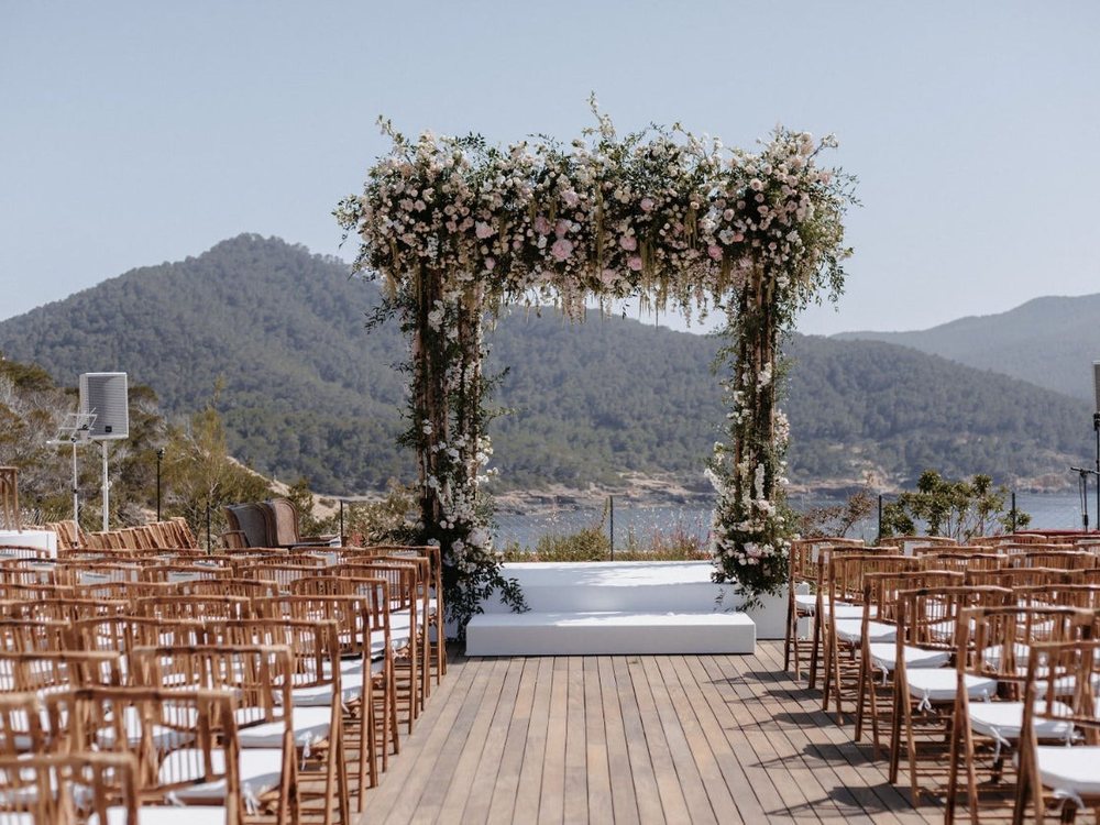 Une cérémonie de mariage installée sur un deck en bois du Six Senses Ibiza, arche fleurie face à la baie
