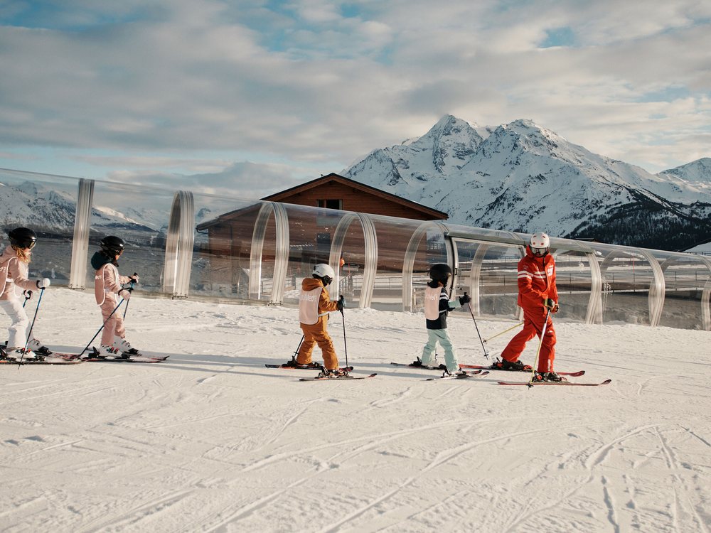 Une file d'enfants en combinaison de ski sur la neige du Club Med Valmorel, face aux sommets de la Tarentaise