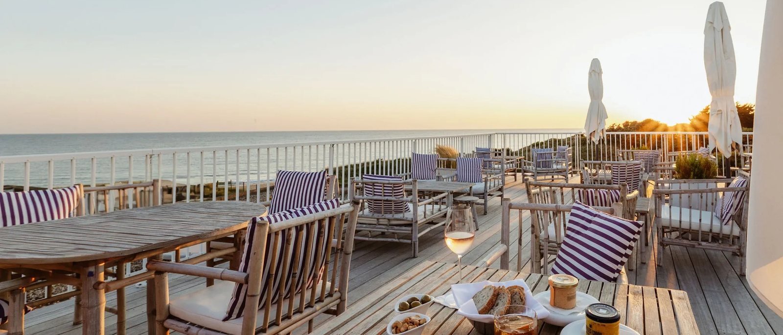 Une terrasse en bois du Grand Large au coucher du soleil, fauteuils rayés, verres et planche apéritive face à l'océan