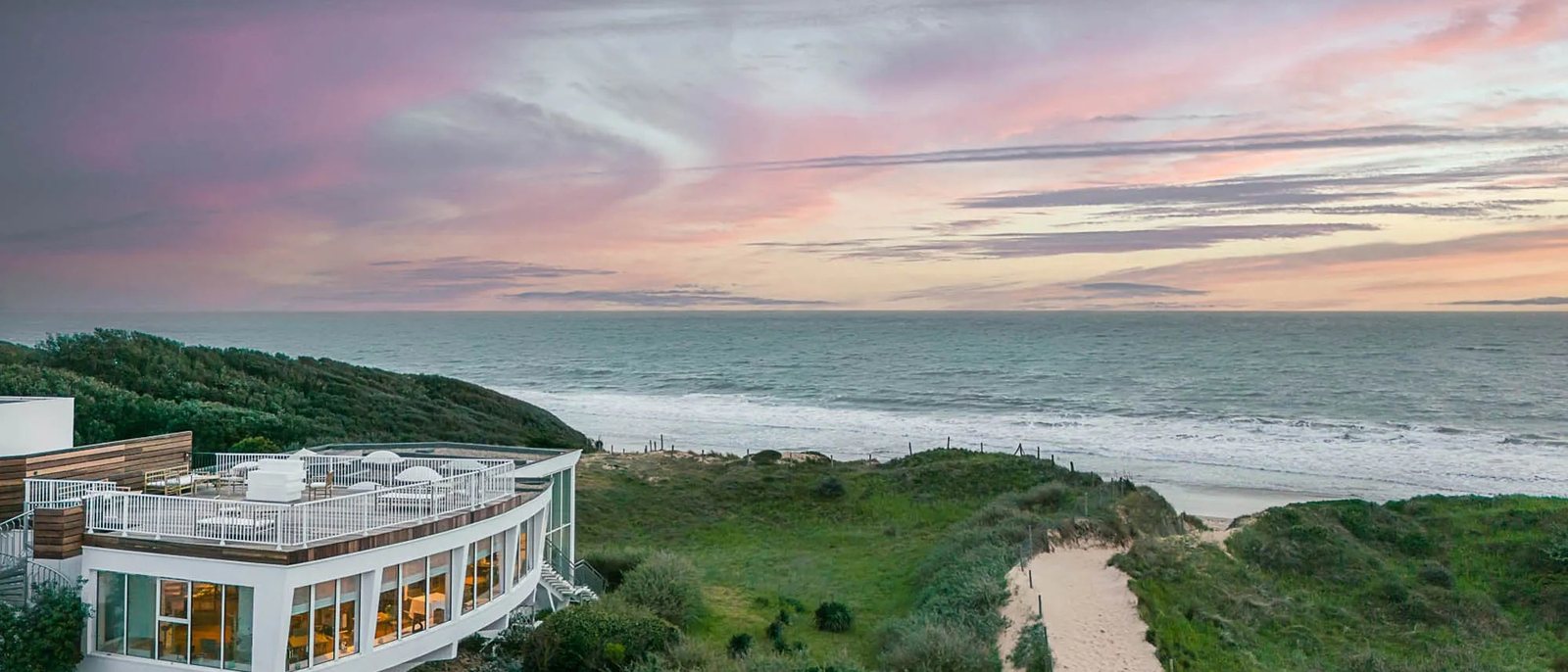 Le Grand Large vu du ciel au coucher du soleil, son aile arrondie et sa terrasse sur le toit, le sentier des dunes et l'océan