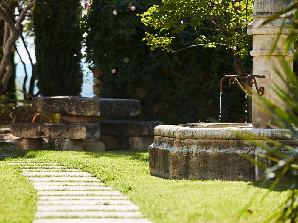 Une fontaine de pierre et une allée dallée dans le jardin du Domaine des Andéols, sous les arbres
