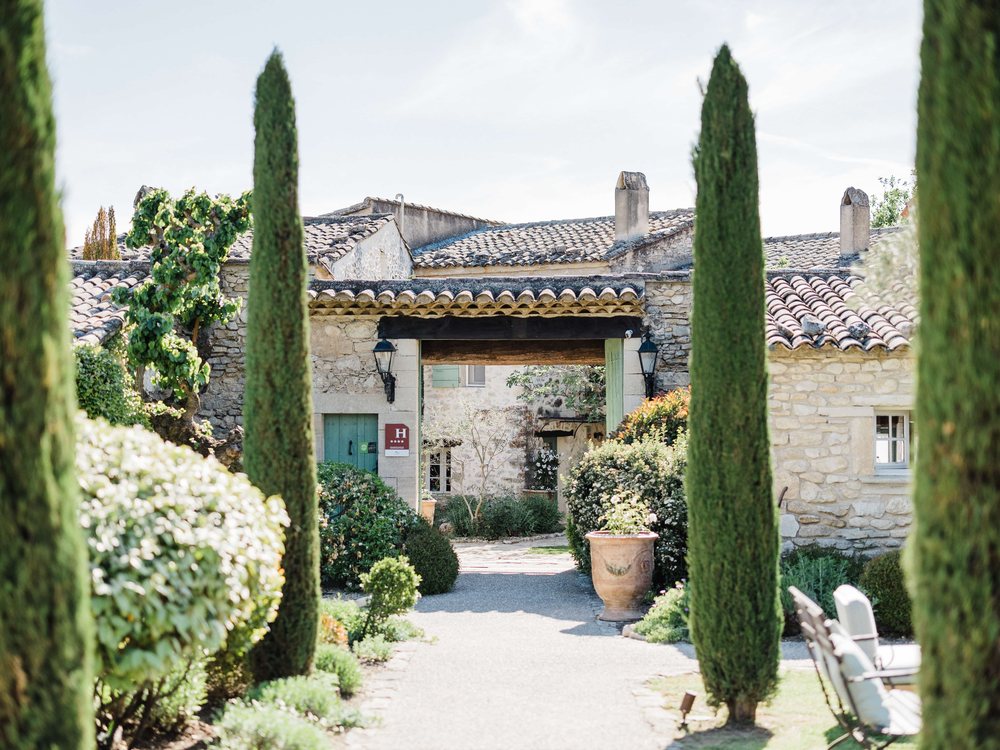 La cour de La Ferme de la Huppe, ses cyprès, ses massifs fleuris et la façade de pierre de l'ancienne ferme