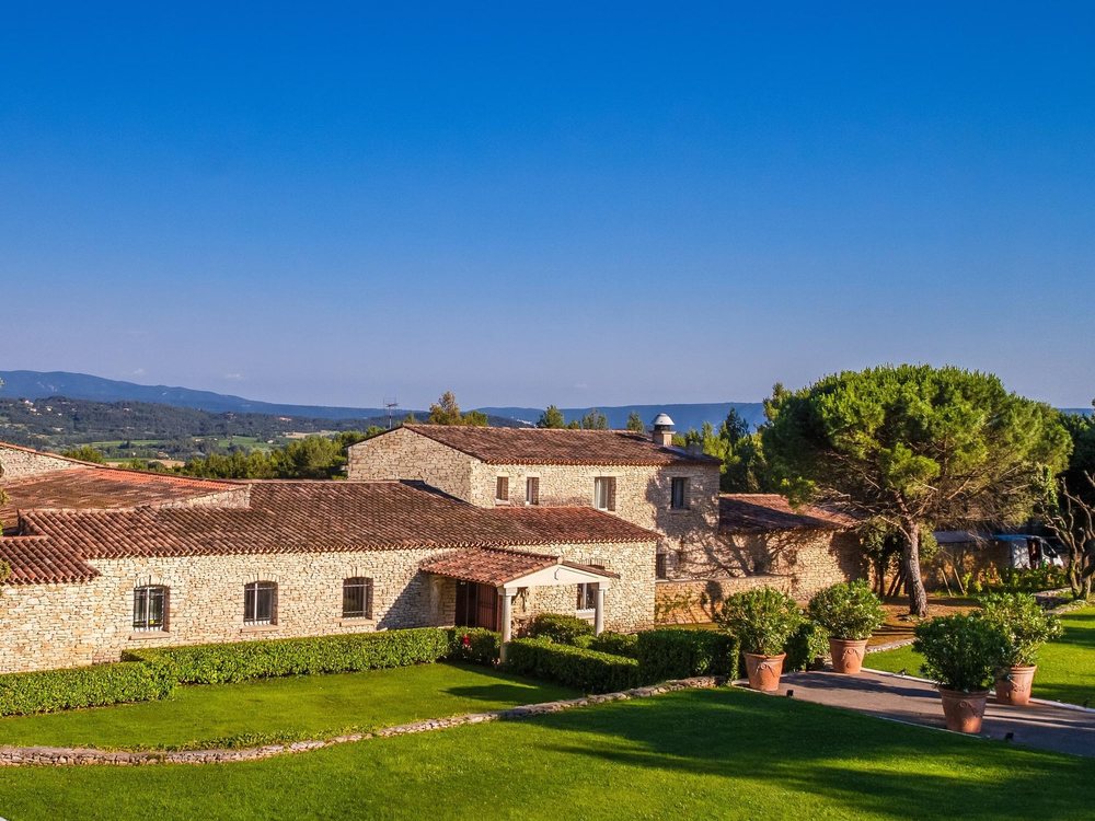 Le Phébus et ses bâtiments de pierre au milieu des pelouses, face aux collines du Luberon sous le ciel bleu