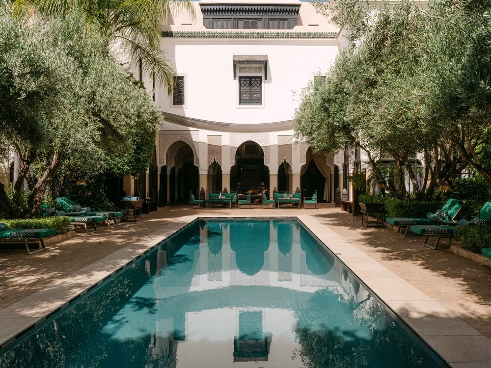 Le patio de La Villa des Orangers, longue piscine bordée d'oliviers devant la façade blanche à arcades