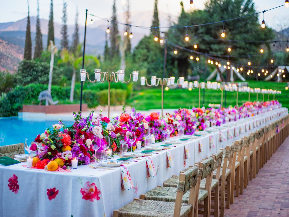 Une longue table de mariage fleurie au bord de la piscine de la Kasbah Tamadot, guirlandes lumineuses et montagnes