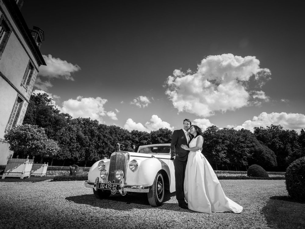 Des mariés devant une voiture ancienne dans la cour du Château de Bourron, en noir et blanc
