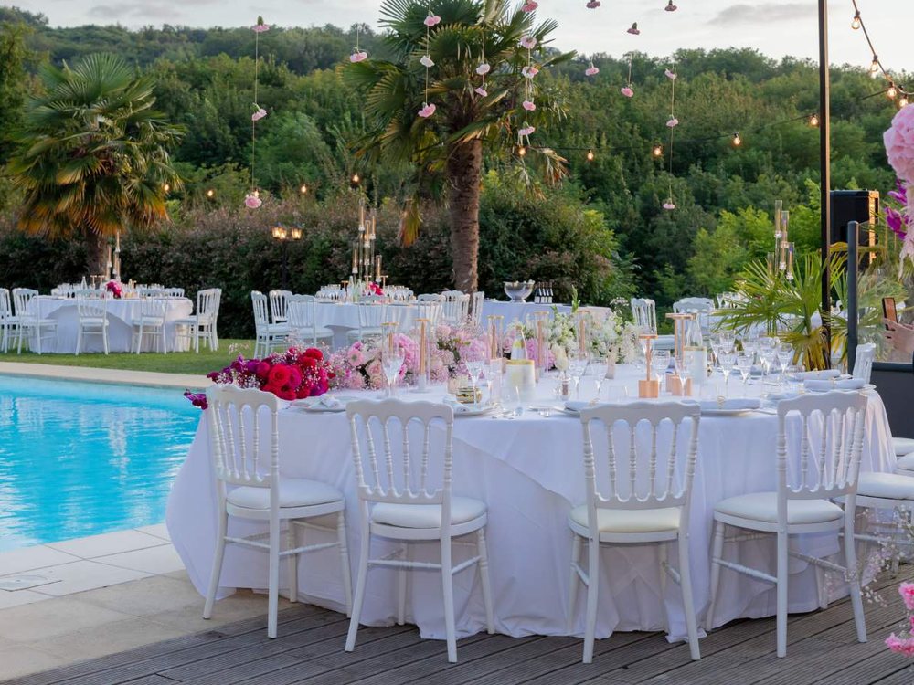 Des tables rondes dressées pour un mariage au bord de la piscine du Château Chapeau Cornu, sous les palmiers