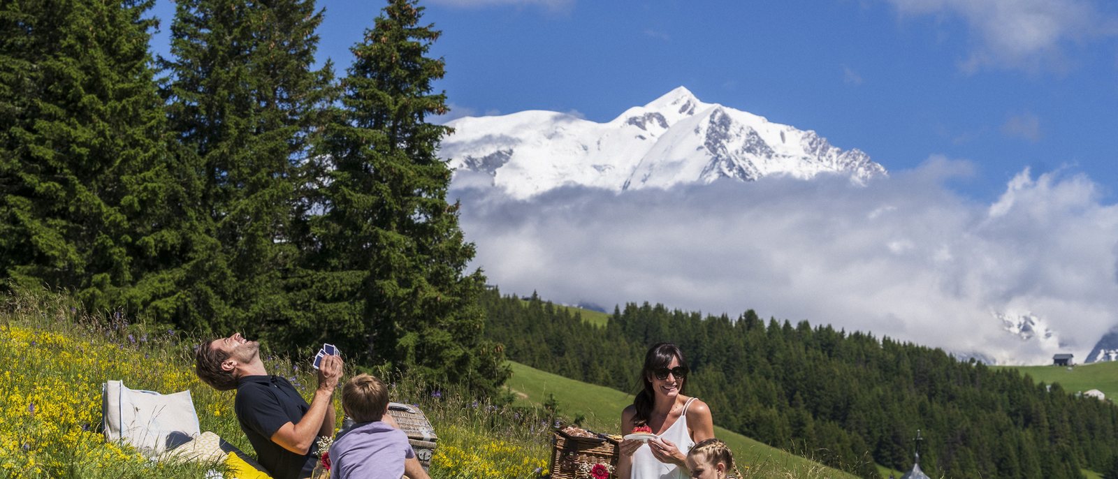 Une famille pique-nique dans un alpage fleuri au-dessus de Megève, face aux sommets enneigés, un jour d'été