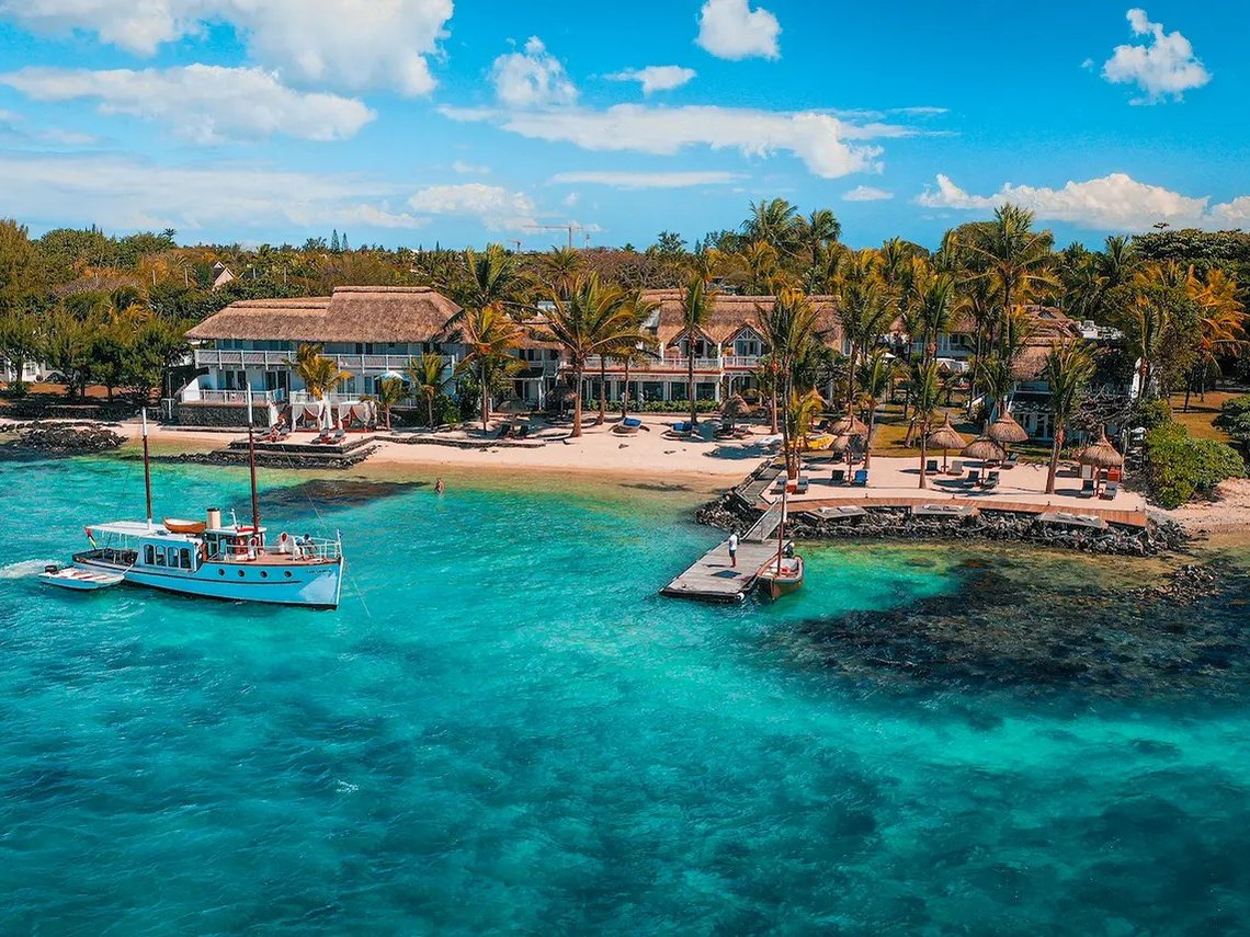 La Maison 20 Degrés Sud vue du ciel, sa plage bordée de cocotiers et un bateau ancien au mouillage sur le lagon de Grand Baie
