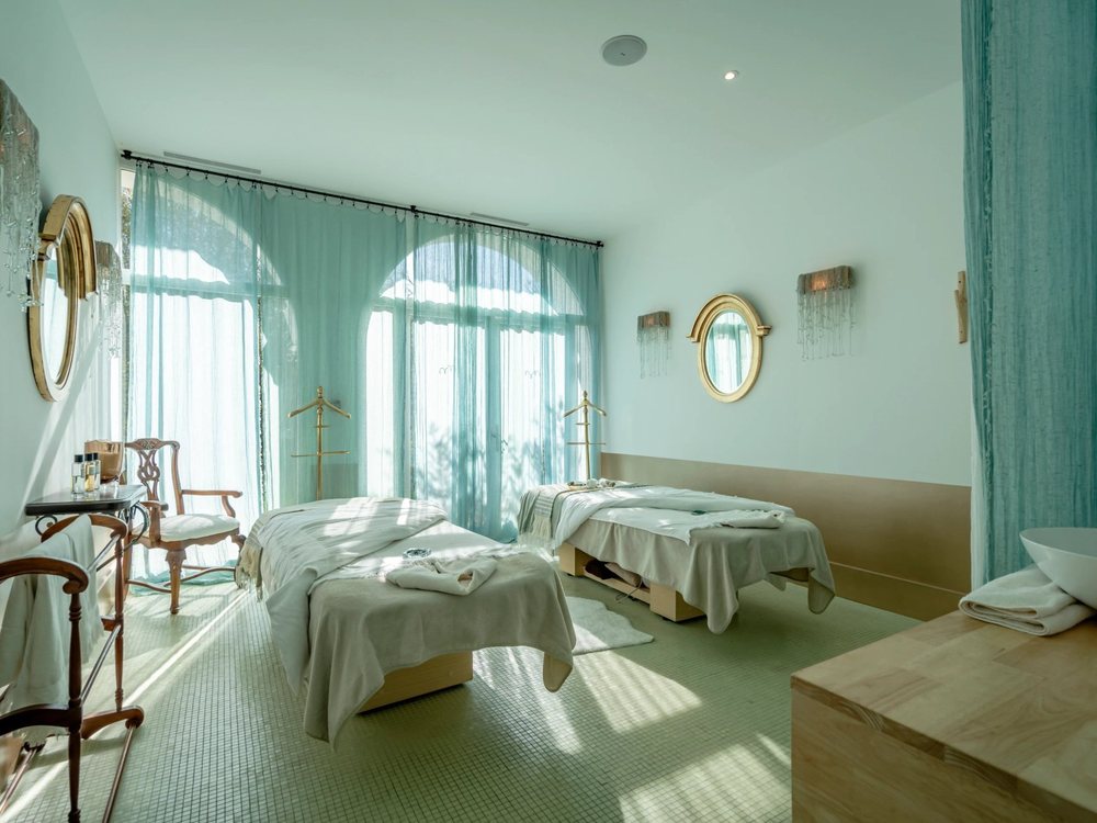 Une cabine de soins en duo du spa de Brindos, ses tables blanches et ses grandes fenêtres voilées de rideaux clairs