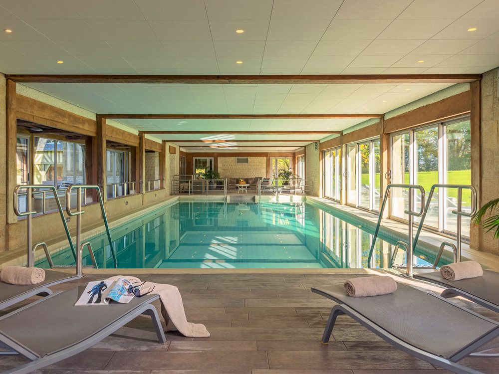 La piscine intérieure chauffée des Manoirs de Tourgéville, ses transats et ses baies vitrées sur le parc