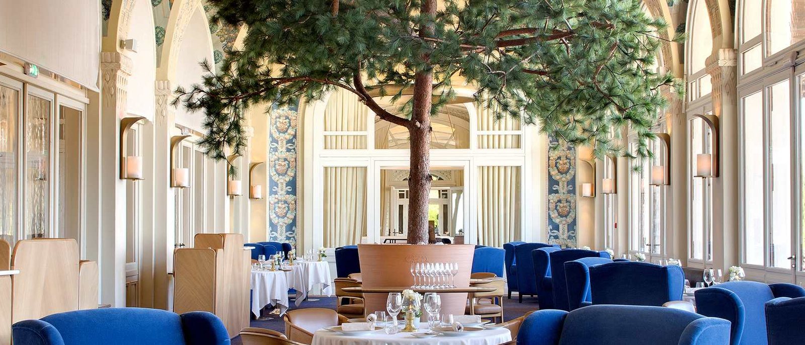 Une salle de restaurant de l'Hôtel Royal, ses fauteuils bleus, ses tables dressées et un arbre sous une verrière