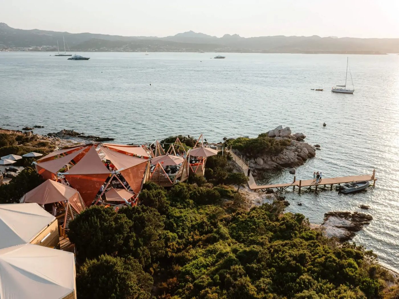 Le beach club du 7Pines Resort Sardinia au coucher du soleil, ses voiles orangées au bord de l'eau, un ponton et le golfe d'Arzachena