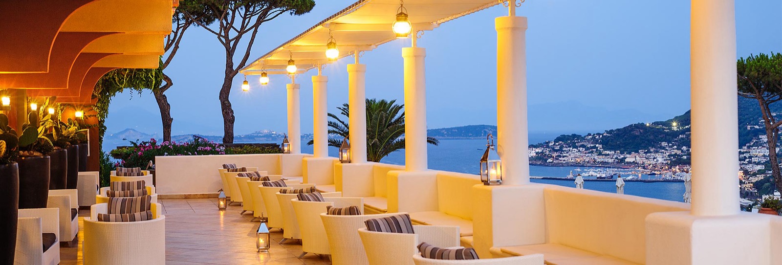 La terrasse du Sunset Lounge du San Montano au crépuscule, colonnes blanches, lanternes et vue sur la baie de Lacco Ameno