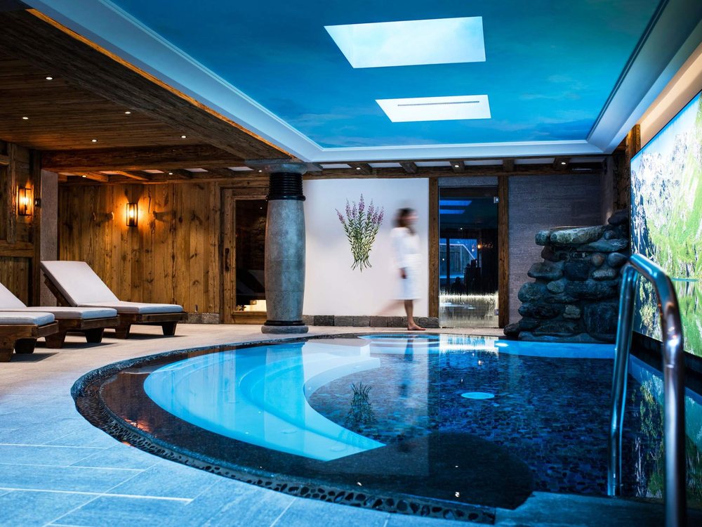 La piscine intérieure du spa La Bèla Vya de La Bouitte, entre pierre et bois, sous un ciel de lumière bleue