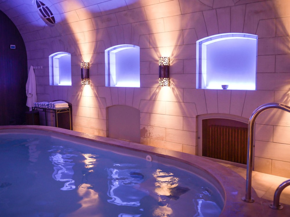 Le bassin du spa du Château d'Artigny sous une voûte de pierre claire, niches éclairées et lumière violette