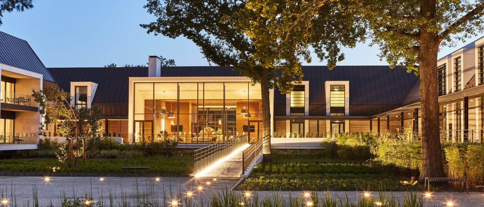 La cour du Domaine des Vanneaux à la tombée du jour, le lobby vitré éclairé sous son toit en pente, un grand arbre et les jardins bordés de lumières