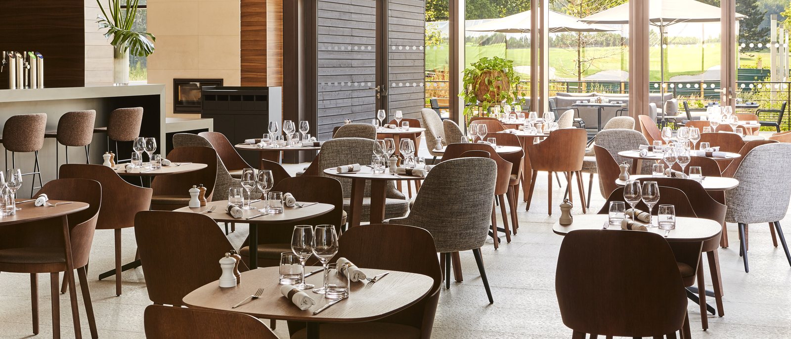 La salle du restaurant Le Piaf, ses tables dressées, ses chaises de cuir brun et ses grandes baies ouvertes sur la terrasse et le golf