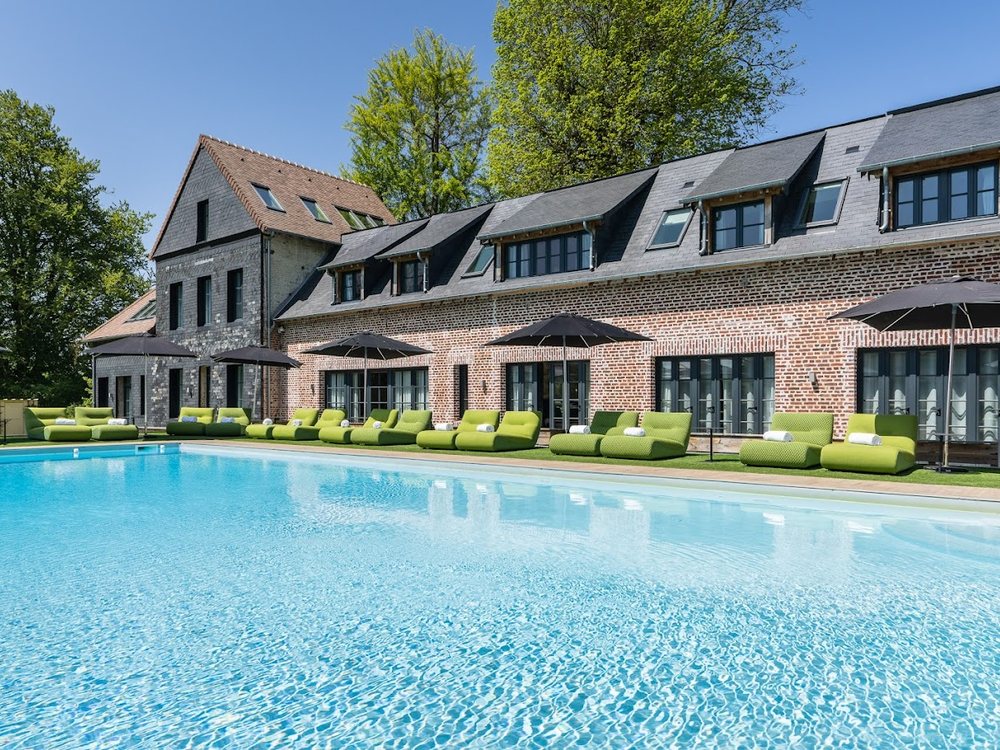 La piscine extérieure des Jardins de Coppélia, ses transats verts devant la façade de briques du manoir